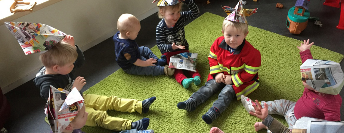 Kinderen zitten samen op het groene kleed, allemaal met een zelfgevouwen krantenhoed op